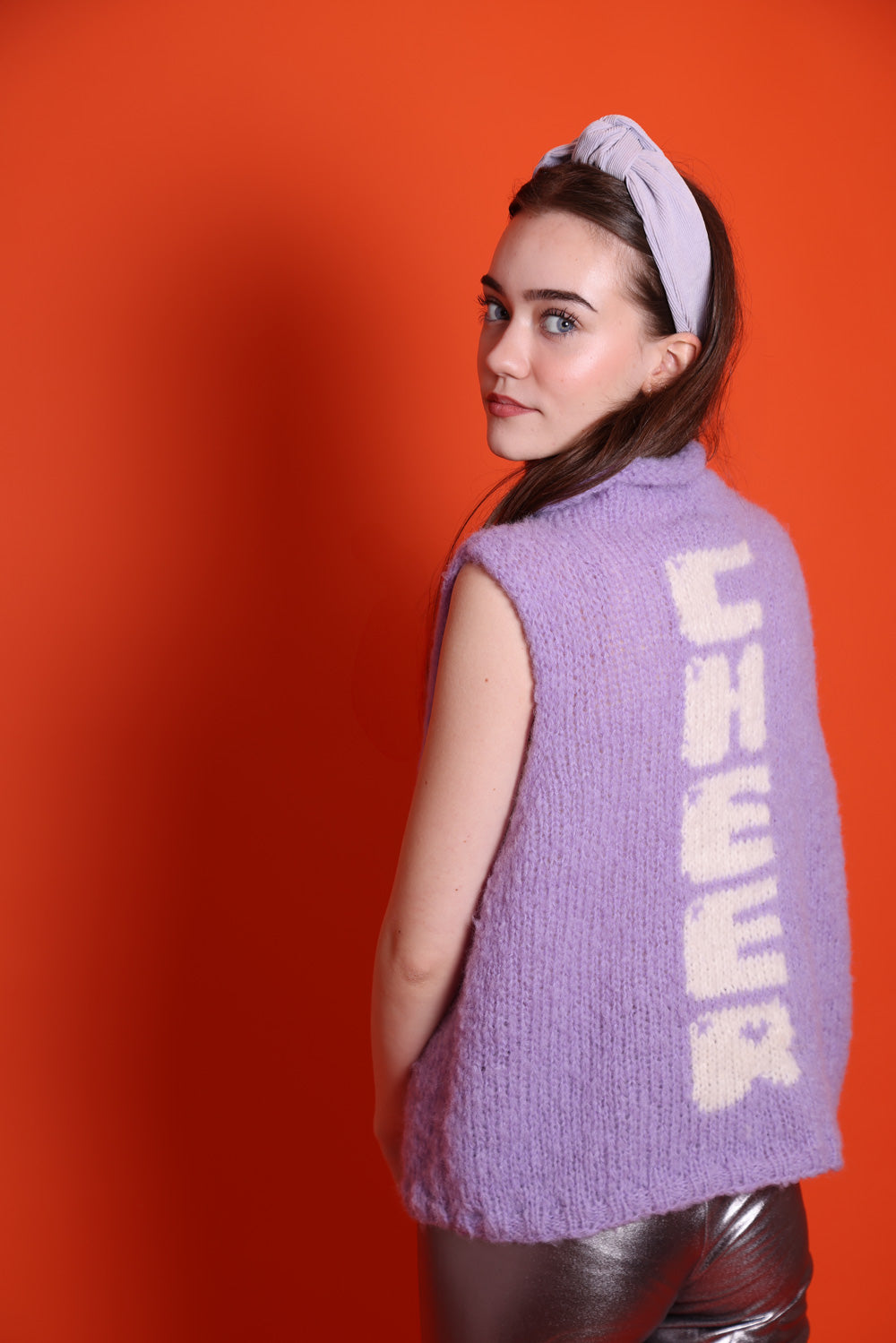 Ein Junges Teeniegirl mit dunklen offenen Haaren, einem lila Haarreif und einem lilafarbenem Wollpullunder steht vor einer orangefarbenen Wand. Sie trägt zudem eine silberne Leggings. Sie ist ein Senior. Auf der Rückseite des Pullunders ist das Wort Cheer eingestrickt. Die Buchstaben laufen untereinander und sind weiß.