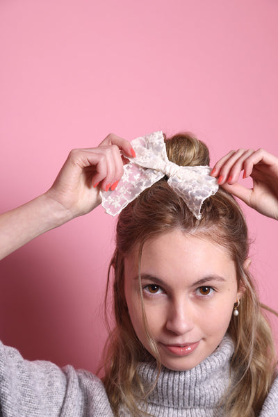 Junge Frau 20 Jahre alt, hat eine Halfbun Frisur mit einer weißen bestickten Schleife auf dem Kopf, die sie mit den Händen richtet. Sie ist im Halbportrait zu sehen, hat dunkelblonde Haare, braune Augen und trägt einen grauen Rollkragenpullover. Sie lächelt und man sieht Perlenohrringe. Ihre Fingernägel sind Corallefarben. Der Hintergrund ist rosa. Verkauft wird die Schleife.