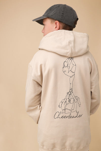 Junge steht mit dem Rücken zum Betrachter. Er trägt eine graue Capi, einen beigen Pullover mit einem großen Backprint mit einer One Line Zeichnung eines Cheerleading Groupstunts und der Aufschrift Cheerleader vor beigem Hintergrund.