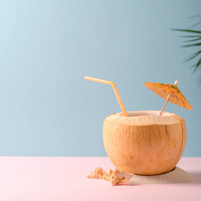 Sommercocktail in Kokussnussschale mit Strohhalm und Papierschirmchen steht auf Sand neben Muschel auf rosafarbenem Untergrund und hellblauem Hintergrund. Am rechten Rand sieht man grüne Blätetr einer Palme.