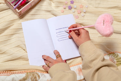 Aufgeschlagenes Cheerleading Journal liegt auf einer Wolldecke. In das Buch schreibt eine Hand mit beigem Pullover etwas mit einem rosa Plüschstift rein.