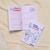 Aufgeschlagenes Cheerleading Journal und eins in geschlossen liegen auf einer Wolldecke. Das Buch hat ein illustriertes Cover mit vielen Cheerleadingsymbolen. Beim aufgeschlagenen Buch sieht man eine Kalenderübersicht in Pastelltönen.