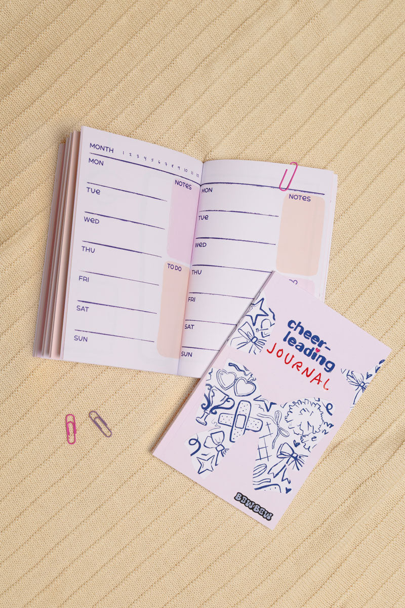 Aufgeschlagenes Cheerleading Journal und eins in geschlossen liegen auf einer Wolldecke. Das Buch hat ein illustriertes Cover mit vielen Cheerleadingsymbolen. Beim aufgeschlagenen Buch sieht man eine Kalenderübersicht in Pastelltönen.