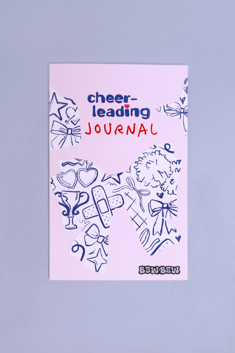 Cheerleading Journal liegt auf einem lila Hintergrund. Das Buch hat ein illustriertes Cover mit vielen Cheerleadingsymbolen.