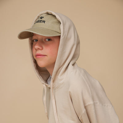 Ein Junge, der im Halbprofil in die Kamera guckt, trägt einen beigen Hoodie, dessen Kapuze auf einer beigen Cap ist. Auf der Cap ist das Motiv: I BOW (Schleifen-Symbol) CHEER. Der Junge schaut freundlich und steht vor einem beigen Hintergrund.