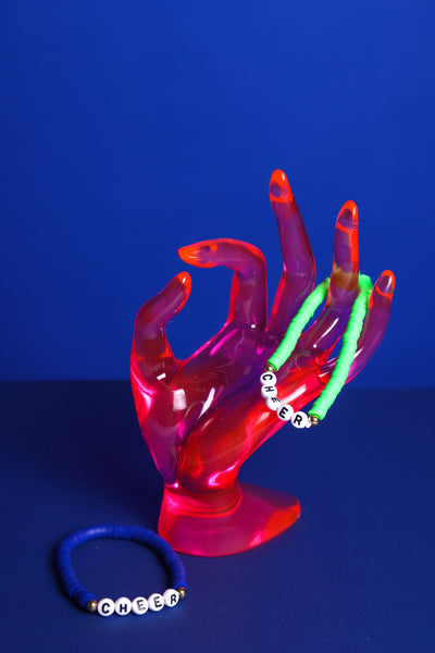 Grünes Perlenarmband mit weißen Perlen mit der Aufschrift Cheer hängt auf einer Acrylglas Hand in durchsichtig pink. Daneben liegt auf dem Boden ein Blaues Perlenarmband mit weißen Perlen und der Aufschrift Cheer. Der Hintergrund ist blau.