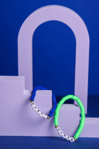 Grünes und blaues Perlenarmband, je mit weißen Perlen mit der Aufschrift Cheer hängne auf einer lilafarbene Treppe Daneben liegt auf dem Dahinter ist ein lilafarbener Torbogen. Der Hintergrund ist blau. 