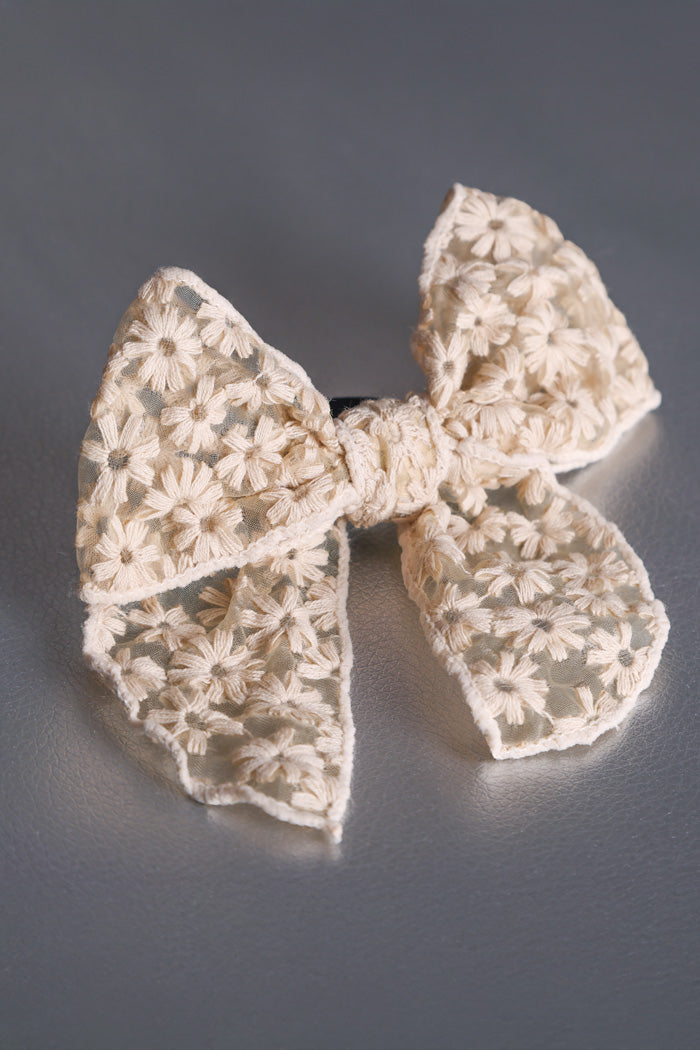 Das Produkt ist eine Beige Haarschleife mit schwarzem Haargummi. Das Schleifenband ist umsümt und auf den durchsichtigen Stoff sind mit beigem Faden Blumen draufgestickt. Der Stoff hat einen Charkter von Spitze. Das Produkt liegt auf einem silbernen Untergrund.