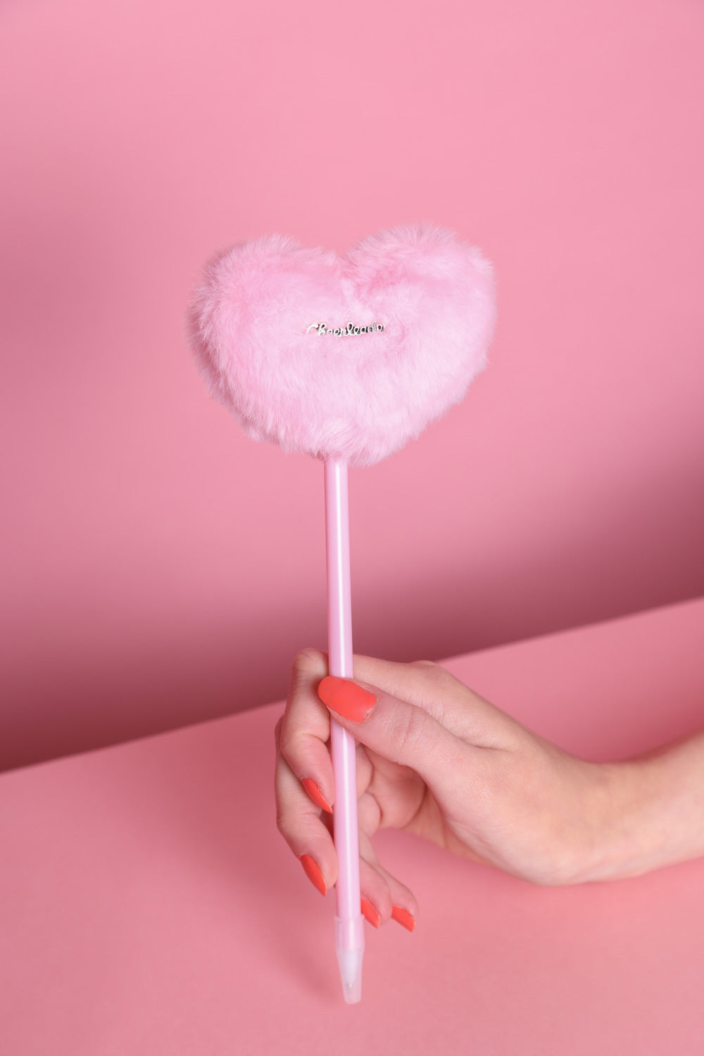 Stift mit Plüschherz auf dem ein silberner Metallaufnäher dran ist mit der Aufschrift: Cheerleader. in der Variante Rosa. Der Stift wird von einer Hand mit orangefarbenem Nagellack gehalten. Der Hintergrund ist rosa.