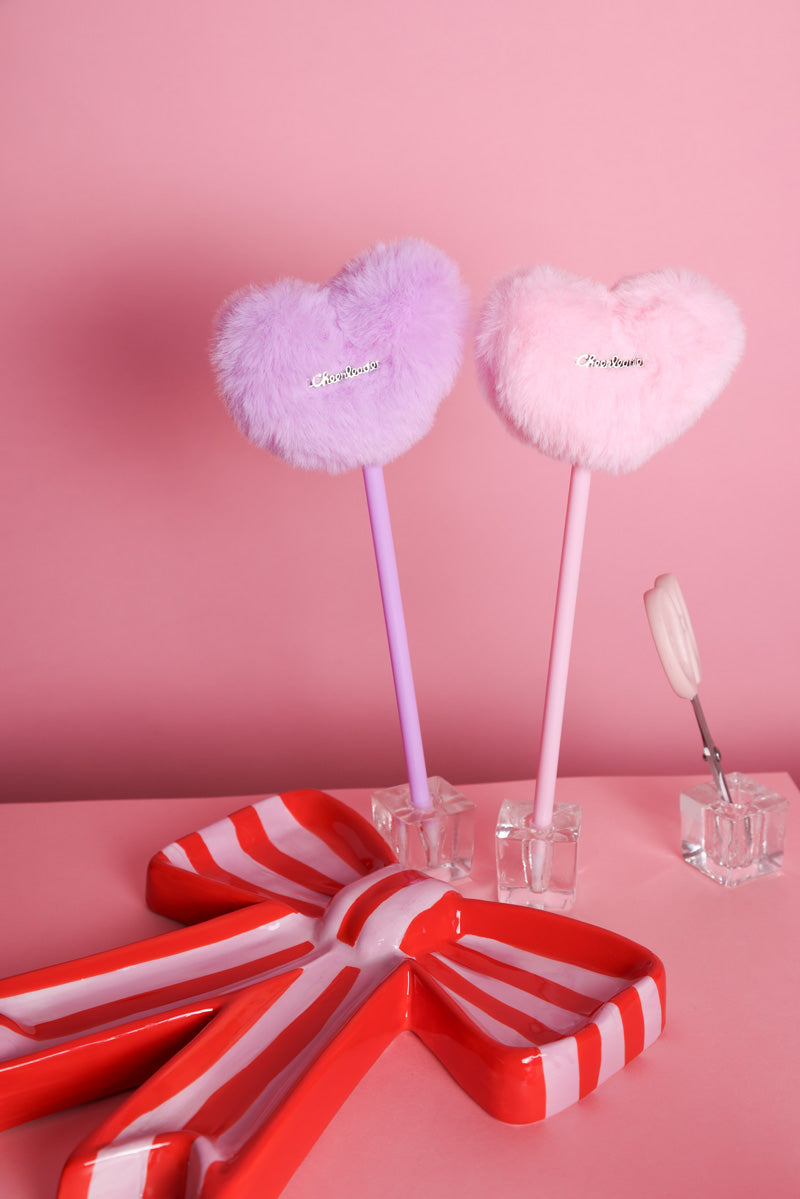 Stifte mit Plüschherz auf dem ein silberner Metallaufnäher dran ist mit der Aufschrift: Cheerleader. Zwei Varianten: Rosa und Lila. Die Stifte stehen hier in kleinen Glas Stiftehaltern auf rosafarbenem Hintergrund. Davor steht eine rosa-rot gestreifte Schale in Schleifenrom. In einem weiteren Glasbehälter steht eine kleine rosafarbene Schere. Der Hintergrund ist ebenfalls Rosa. Verkauft werden die Cheerleader Plüschstifte.