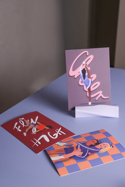Postkartenset. Zwei der drei Postkarten liegen auf einem fliederfarbenem Tisch. Eine steht in einem Postkartenhalter in. Im Hintergrund sieht man in der Unschärfe einen Raum. 