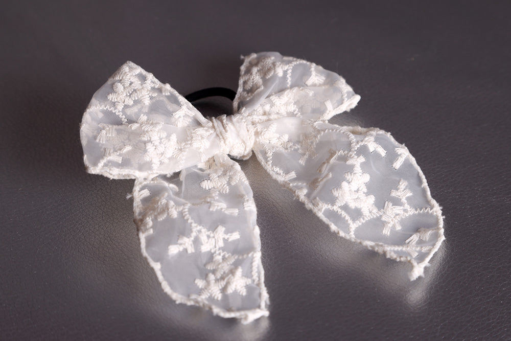 Das Produkt ist eine offwhite-farbene Haarschleife mit schwarzem Haargummi. Das Schleifenband ist umsäumt und auf den durchsichtigen Stoff sind mit offwhite farbenem Faden florale Muster draufgestickt. Der Stoff hat einen Charkter von Spitze. Das Produkt liegt auf einem silbernen Untergrund.