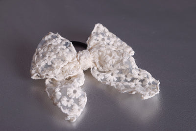 Das Produkt ist eine creme-farbene Haarschleife mit schwarzem Haargummi. Das Schleifenband ist umsäumt und auf den durchsichtigen Stoff sind mit creme-farbenem Faden Blumen-Muster draufgestickt. Der Stoff hat einen Charkter von Spitze. Das Produkt liegt auf einem silbernen Untergrund.