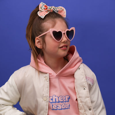 Ein kleines Mädchen mit mittelblonden Haaren, einer rosa Herzbrille, einer bestickten Schleife und einem rosafarbenem Hoodie mit der Aufschrift: Cheerleader in Pixelart in lila,rosa,weiß, steht vor einer blauen Wand .Sie trägt eine beige Jacke. Sie ist ein Peewee und man sieht sie im Halbprofil. Sie lächelt und man erkennt ihre süßen Zähne mit Zahnlücke.
