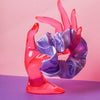 Ein lilafarbenes Scrunchie ist um den Finger, einer pinke Acrylglas-Hand gebunden, die auf lilanem Untergrund und vor rosanem Hintergrund steht.  