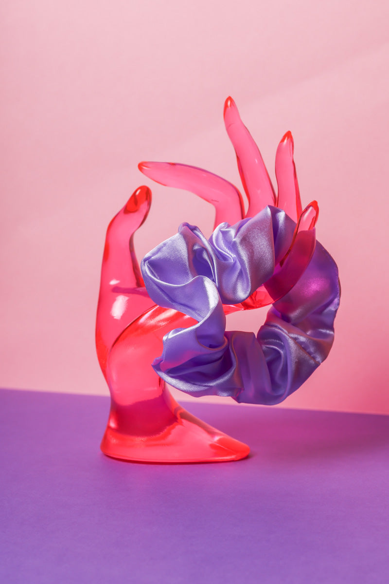 Ein lilafarbenes Scrunchie ist um den Finger, einer pinke Acrylglas-Hand gebunden, die auf lilanem Untergrund und vor rosanem Hintergrund steht.  
