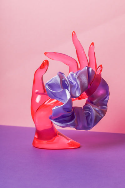 Ein lilafarbenes Scrunchie ist um den Finger, einer pinke Acrylglas-Hand gebunden, die auf lilanem Untergrund und vor rosanem Hintergrund steht.  
