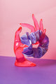 Ein lilafarbenes Scrunchie ist um den Finger, einer pinke Acrylglas-Hand gebunden, die auf lilanem Untergrund und vor rosanem Hintergrund steht.  