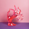Ein corallefarbenes Scrunchie ist um den Finger, einer pinke Acrylglas-Hand gebunden, die auf lilanem Untergrund und vor rosanem Hintergrund steht.  