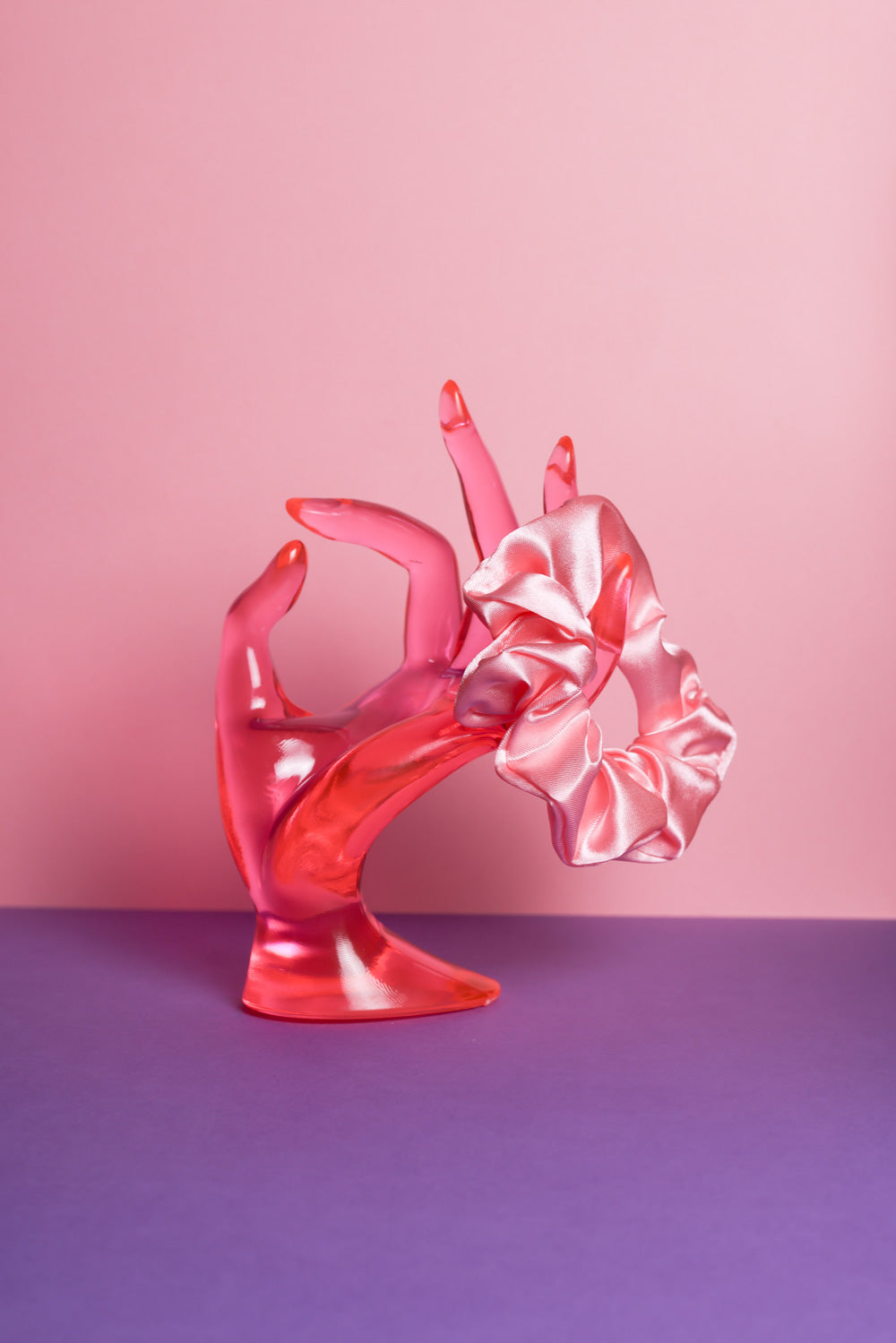 Ein corallefarbenes Scrunchie ist um den Finger, einer pinke Acrylglas-Hand gebunden, die auf lilanem Untergrund und vor rosanem Hintergrund steht.  