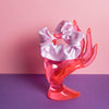 Ein hell lilafarbenes Scrunchie ist um den Finger, einer pinke Acrylglas-Hand gebunden, die auf lilanem Untergrund und vor rosanem Hintergrund steht.  
