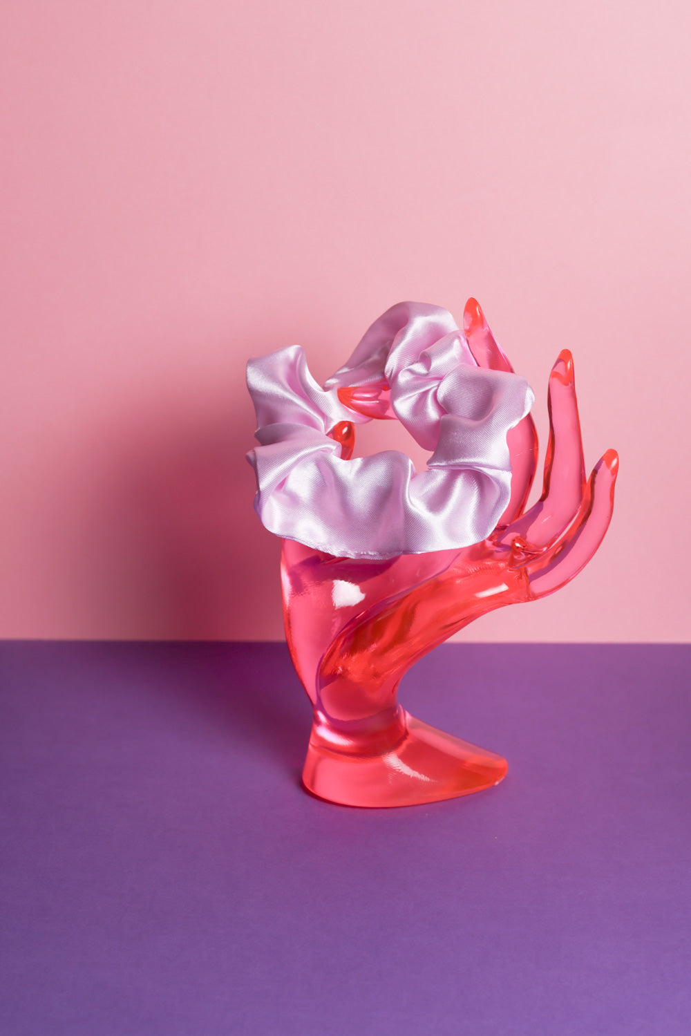 Ein hell lilafarbenes Scrunchie ist um den Finger, einer pinke Acrylglas-Hand gebunden, die auf lilanem Untergrund und vor rosanem Hintergrund steht.  
