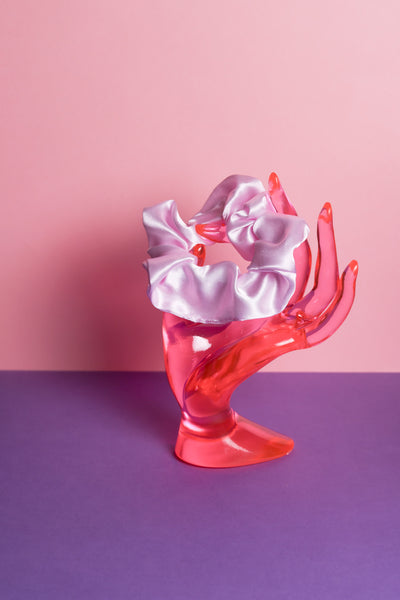 Ein hell lilafarbenes Scrunchie ist um den Finger, einer pinke Acrylglas-Hand gebunden, die auf lilanem Untergrund und vor rosanem Hintergrund steht.  