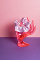 Ein hell lilafarbenes Scrunchie ist um den Finger, einer pinke Acrylglas-Hand gebunden, die auf lilanem Untergrund und vor rosanem Hintergrund steht.  
