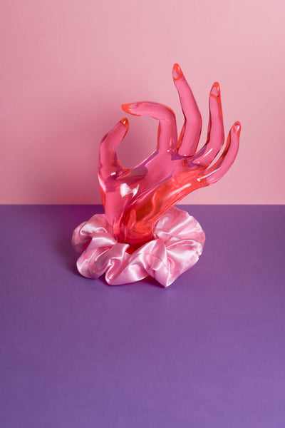 Ein rosafarbenes Scrunchie ist um eine pinke Acrylglas-Hand gebunden, die auf lilanem Untergrund und vor rosanem Hintergrund steht. 