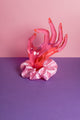 Ein rosafarbenes Scrunchie ist um eine pinke Acrylglas-Hand gebunden, die auf lilanem Untergrund und vor rosanem Hintergrund steht. 