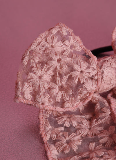 Haarschleife rosefarben, mit eingesticktem Blumenmuster auf Organzastoff mit schwarzem Haargummi liegt in einer Detailaufnahme auf rosanem strukturiertem Untergrund.