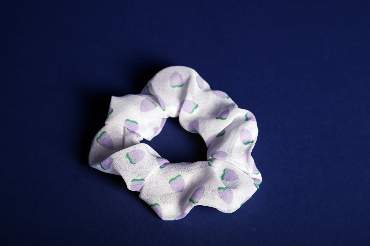 Weißes Scrunchie mit drauf gedruckten, lilafarbenen Erdbeeren mit grünen Blättern, liegt auf einem blauen Untergrund.