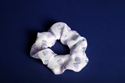 Weißes Scrunchie mit drauf gedruckten, lilafarbenen Erdbeeren mit grünen Blättern, liegt auf einem blauen Untergrund.