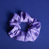 Lilafarbenes Scrunchie mit dunkellilafarbenen Blumen und grüner Mitte liegt auf blauen Untergrund.