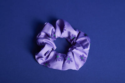 Lilafarbenes Scrunchie mit dunkellilafarbenen Blumen und grüner Mitte liegt auf blauen Untergrund.