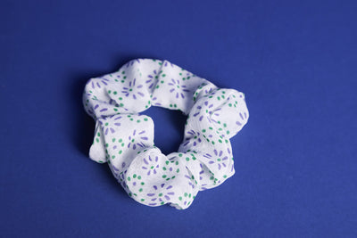 Weißes Scrunchie mit lila - grün getupften floralen Mustern liegt auf einem blauen Untergrund.