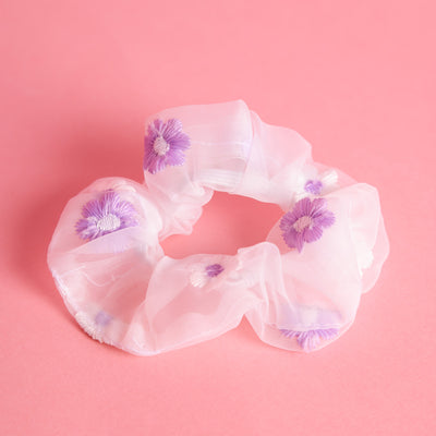 Weißes Scrunchie aus Organzastoff mit weiß-lila bestickten Blumen liegt auf einem rosa farbenem Untergund.