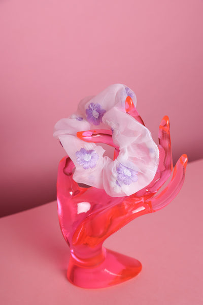 weißes Organzascrunchie mit lila weiß gestickten Blumen drauf, wird von einer pinken Acrylglashand gehalten, die auf rosa Untergrund und vor rosa Hintergrund steht. 