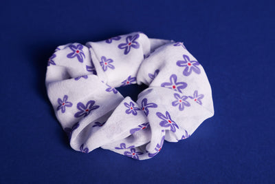 Weißes Scrunchie mit lilafarbenen Blumen und roter Mitte liegt auf einem blauen Untergrund.