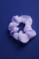 Ein helllilafarbenes Scrunchie aus Organzastoff liegt auf einem blauen Untergrund.