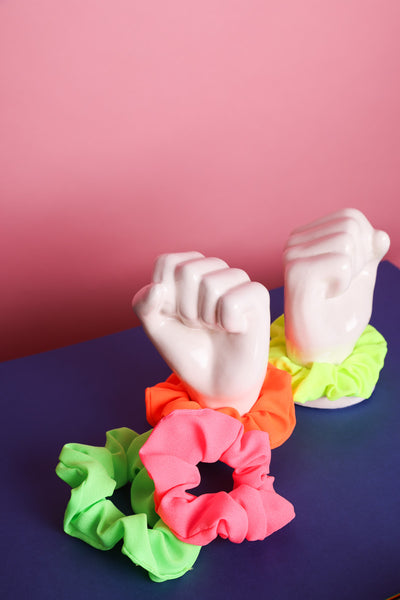 Vier Scunchies aus Stoff in Neongrün, Neonpink auf blauem Untergrund liegend. Dahinter stehen zwei Porzellanhände. Um die eine ist ein Neonorangenes Scrunchie gebunden und um die andere eins in Neongelb. Der Hintergrund ist rosa.