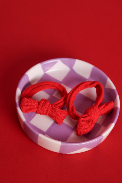 Zwei rote, elastische Haargummis mit integrierter Schleife liegen in einem lila,weiß karierten Porzellanschälchen auf rotem Hintergrund.