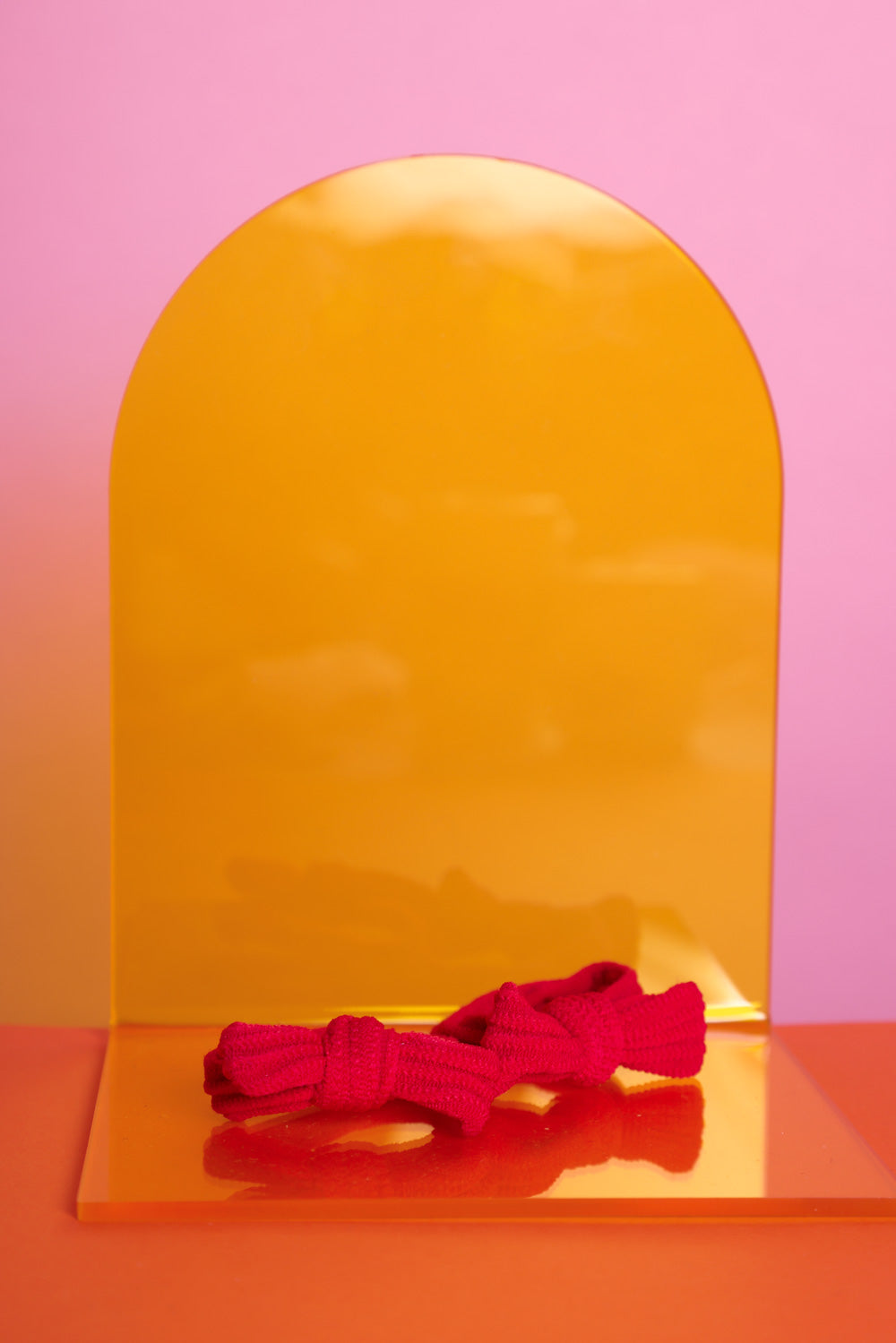 rote, elastische Haargummis in rot, als Doppelpack, liegen auf einem gelben Acrylglasständer auf orangefarbenem Untergrund mit rosafarbenem Hintergrund.