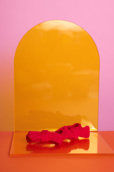 rote, elastische Haargummis in rot, als Doppelpack, liegen auf einem gelben Acrylglasständer auf orangefarbenem Untergrund mit rosafarbenem Hintergrund.