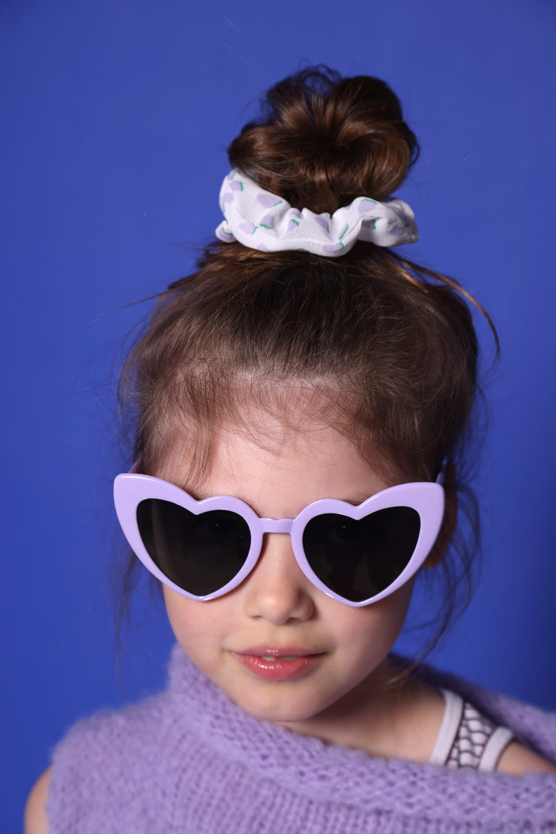 Ein kleines Mädchen mit lilafarbenem Wollpullunder steht vor einer blauen Wand. Sie trägt eine lilafarbene Herzbrille mit dunklen Gläsern. Sie lächelt. Ihre mittelblonden Haare sind im Dutt und sie trägt ein weißes Scrunchie mit lila-grünem Motiv. 