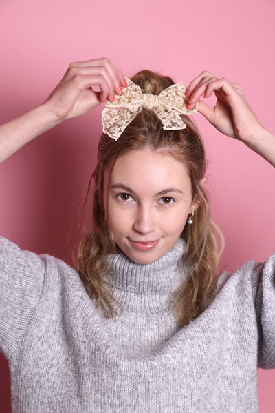 Junge Frau 20 Jahre alt, hat eine Halfbun Frisur mit einer beigen Haarschleife auf dem Kopf, die sie mit den Händen richtet. Sie ist im Halbportrait zu sehen, hat dunkelblonde Haare, braune Augen und trägt einen grauen Rollkragenpullover. Der Hintergrund ist rosa. Verkauft wird die Schleife.