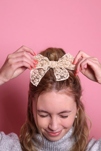 Junge Frau 20 Jahre alt, hat eine Halfbun Frisur mit einer beigen Haarschleife auf dem Kopf, die sie mit den Händen richtet. Sie ist im Portrait zu sehen, hat dunkelblonde Haare, zugekniffene Augen, lächelt und trägt einen grauen Rollkragenpullover und Perlenohrringe. Ihr Nagellack ist Corallefarben. Der Hintergrund ist rosa. Verkauft wird die Schleife.