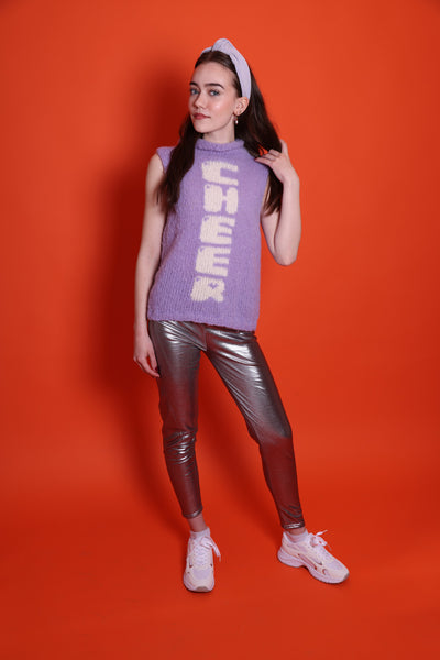 Ein Junges Teeniegirl mit dunklen offenen Haaren, einem lila Haarreif und einem lilafarbenem Wollpullunder steht vor einer orangefarbenen Wand. Sie ist ein Senior. Auf der Vorderseite des Pullunders ist das Wort Cheer eingestrickt. Die Buchstaben laufen untereinander und sind weiß. Sie trägt eine silberne Leggings und weiß-silberne Schuhe. Ihr Gesicht ist im Halbprofil.
