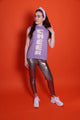 Ein Junges Teeniegirl mit dunklen offenen Haaren, einem lila Haarreif und einem lilafarbenem Wollpullunder steht vor einer orangefarbenen Wand. Sie ist ein Senior. Auf der Vorderseite des Pullunders ist das Wort Cheer eingestrickt. Die Buchstaben laufen untereinander und sind weiß. Sie trägt eine silberne Leggings und weiß-silberne Schuhe. Ihr Gesicht ist im Halbprofil.