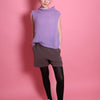 Ein Junges Teeniegirl mit dunklen, zum Scrunchie und einem lilafarbenem Wollpullunder steht vor einer rosa Wand. Sie ist ein Senior. Sie trägt eine graue Short und eine schwarze Strumpfhose und weiße Schuhe. Ihre Hände sind in den Hosentaschen. Sie steht mit frontal zum Betrachter.